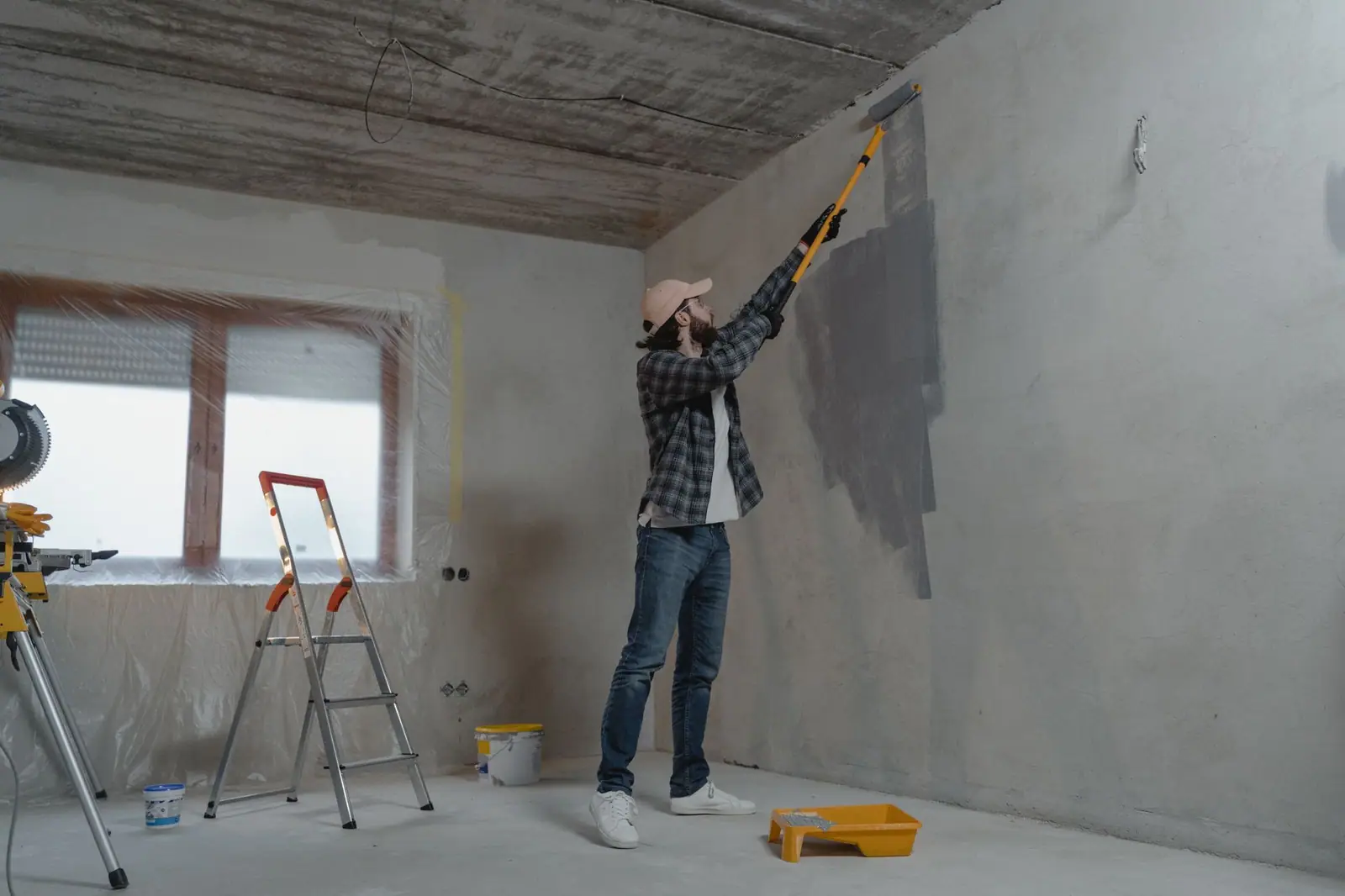 Painter using an extension roller on a gray interior wall with a stepladder nearby