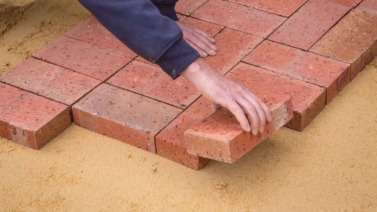 Paver patio cost: hands setting red clay pavers on a sand bed