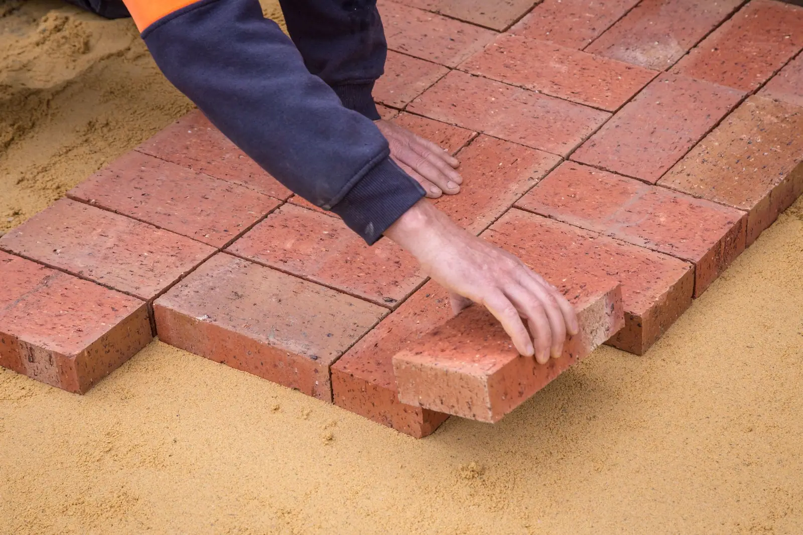 Hands setting red clay pavers in a running-bond pattern on a sand bed