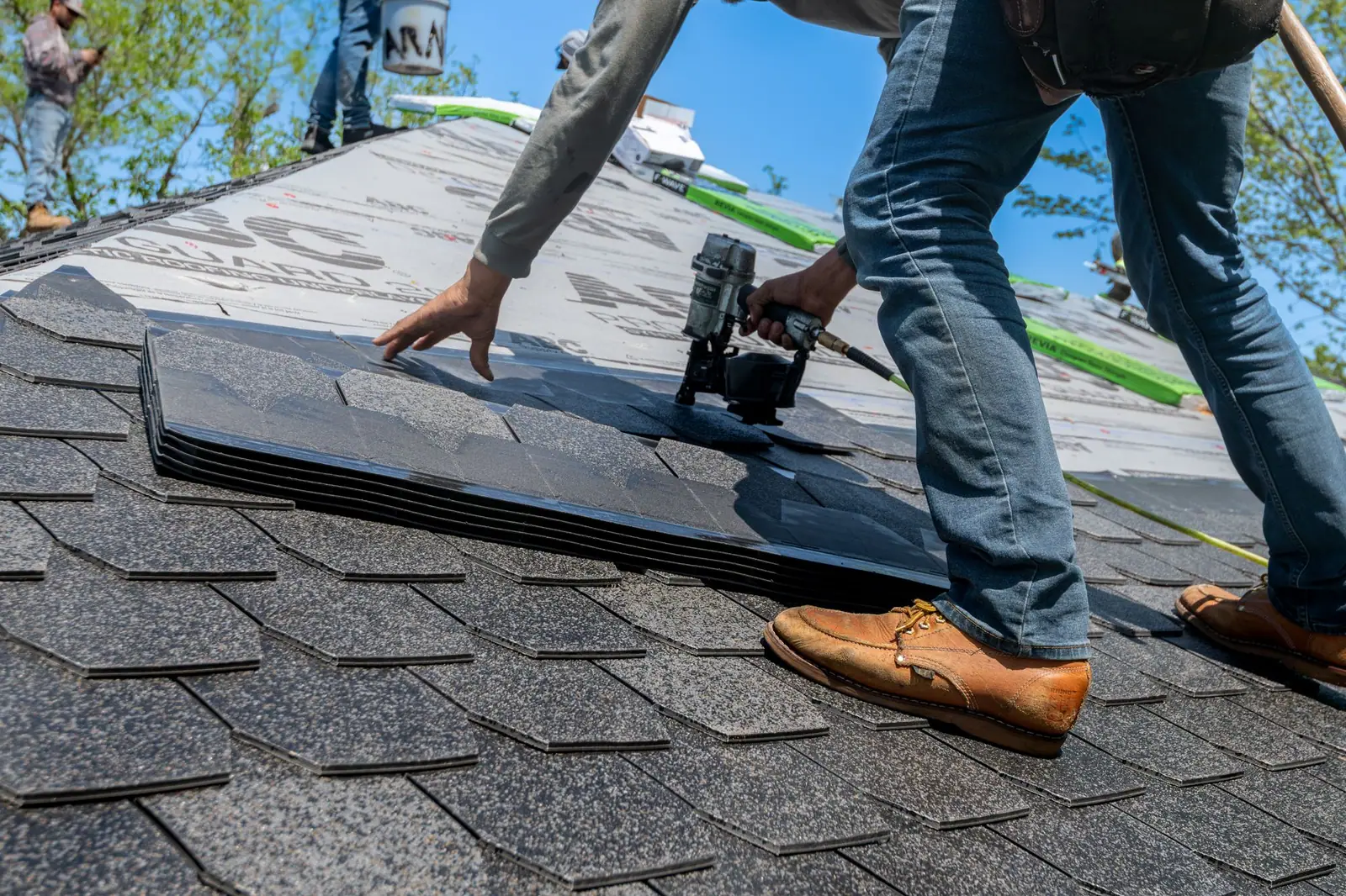 Roofer kneeling on a pitched roof fastening dark asphalt shingles with a nail gun